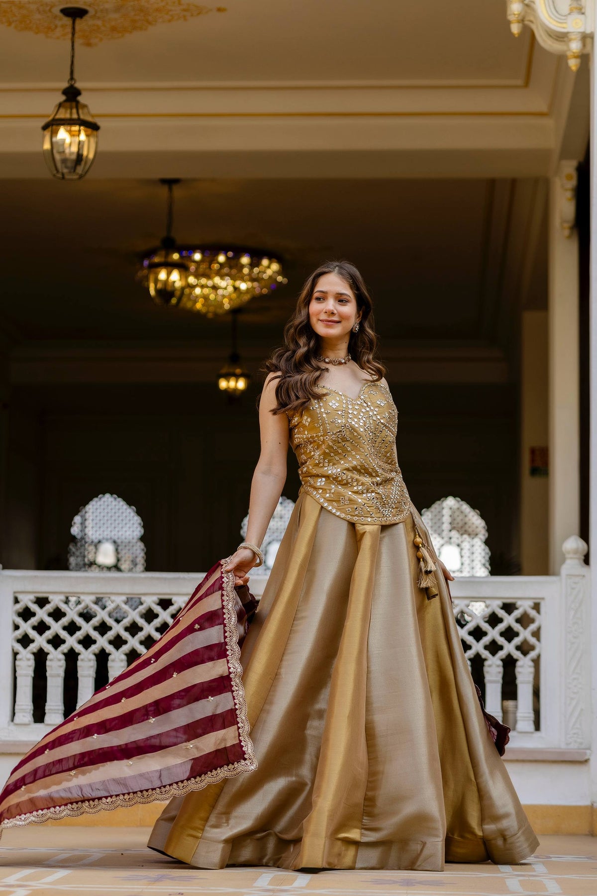 Standing full-length pose of model holding a maroon striped dupatta with scallop trim, paired with a gold flared lehenga
