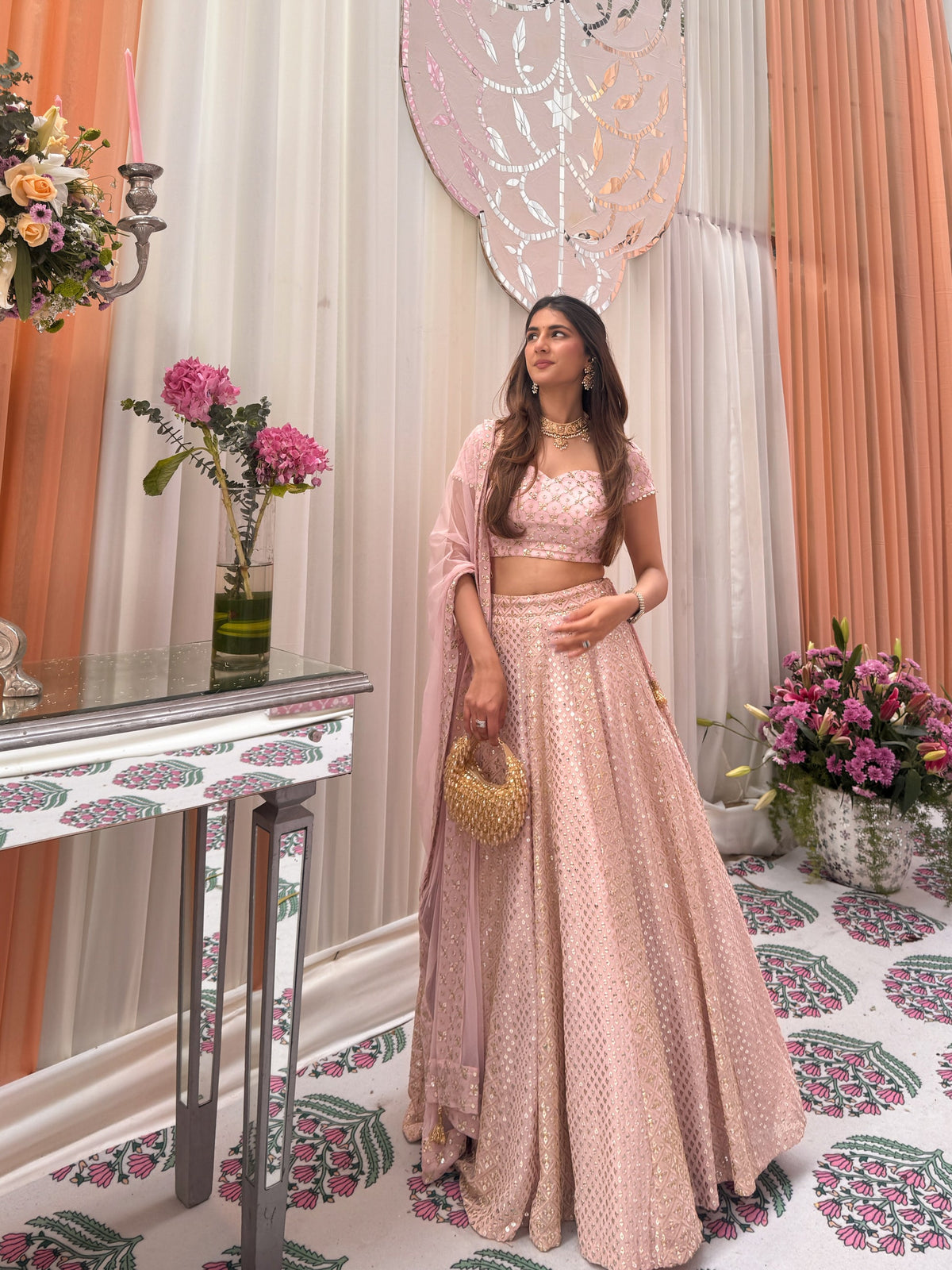 Side-angle full-length shot of a model wearing a baby-pink Chikankari embroidered lehenga with a beaded gold potli bag