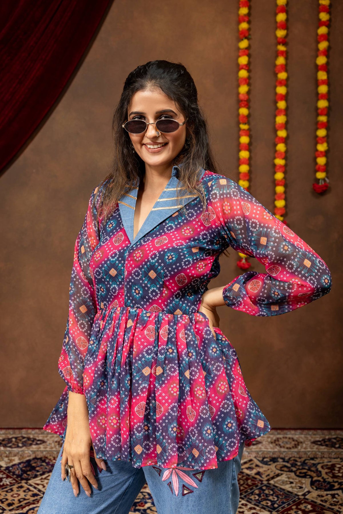 Mid-shot view of model wearing pink and blue printed bandhani peplum top with denim notch lapel collar and sunglasses