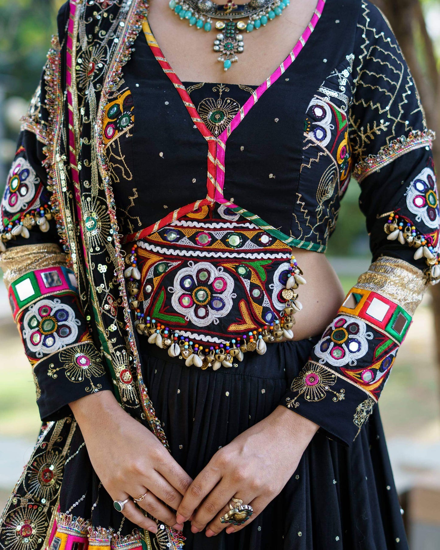 Close-up of black choli featuring central floral mirror patch, colorful piping trim, and hanging cowrie shell tassels at the hem