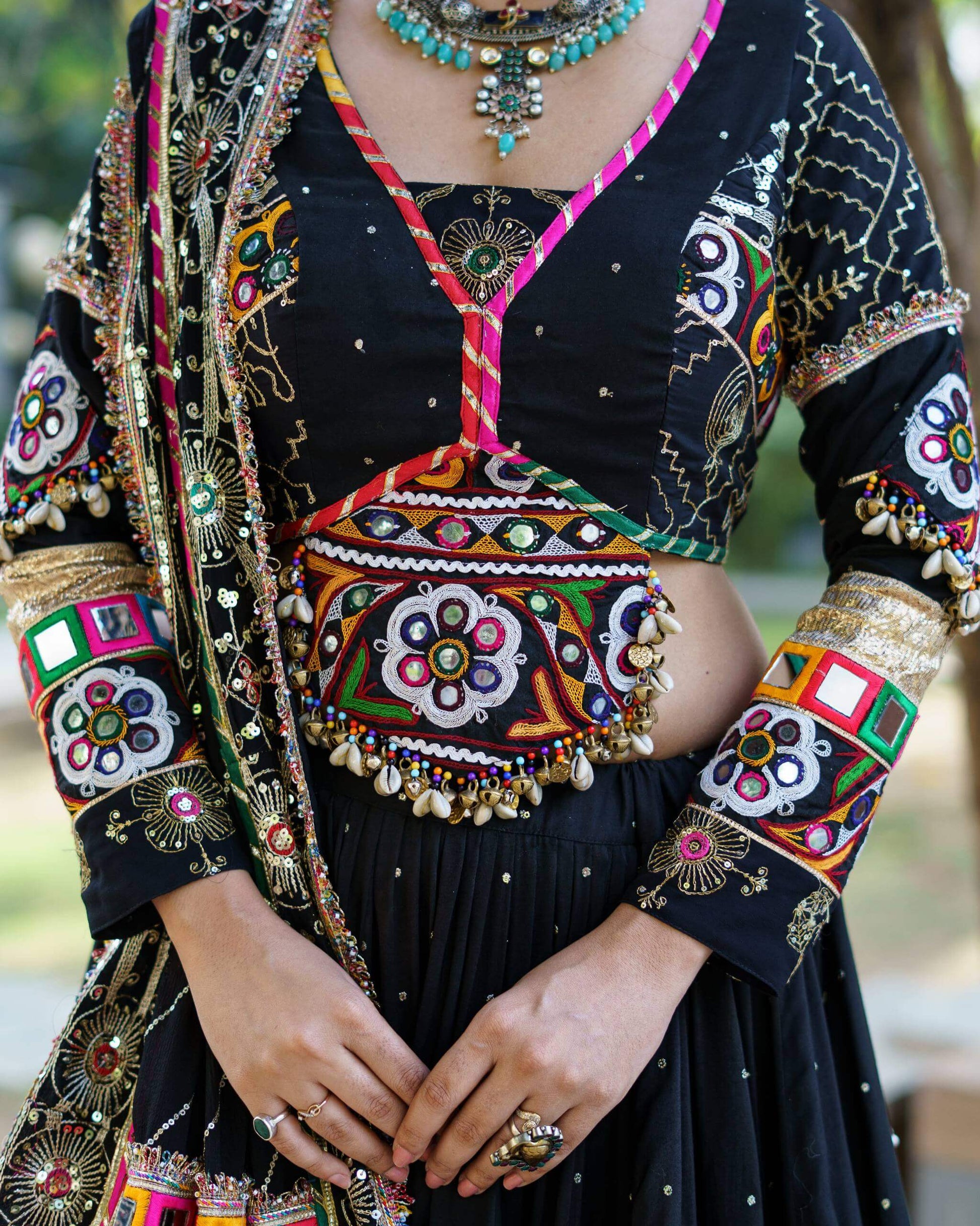 Close-up of black choli featuring central floral mirror patch, colorful piping trim, and hanging cowrie shell tassels at the hem