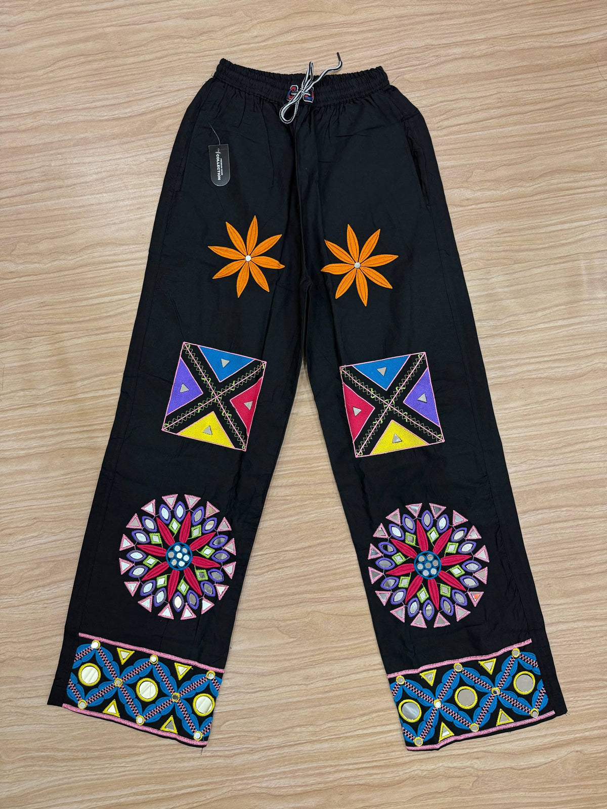 Flat lay view of black palazzo pants featuring embroidered floral, geometric, and mirror-work patchwork designs.