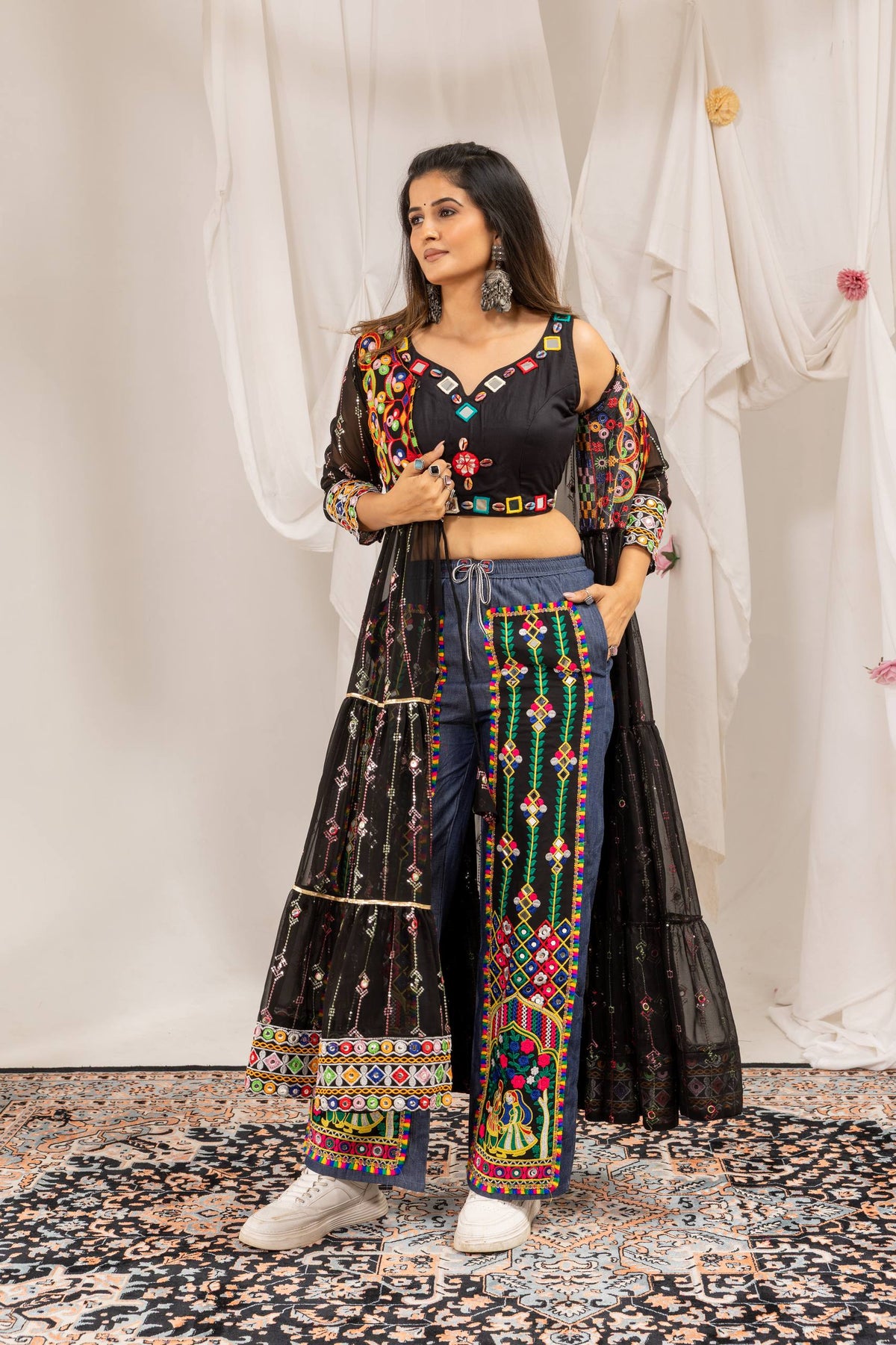 Full-length three-quarter view of model wearing a black mirror-work crop top, tiered sheer shrug, and embroidered denim pants