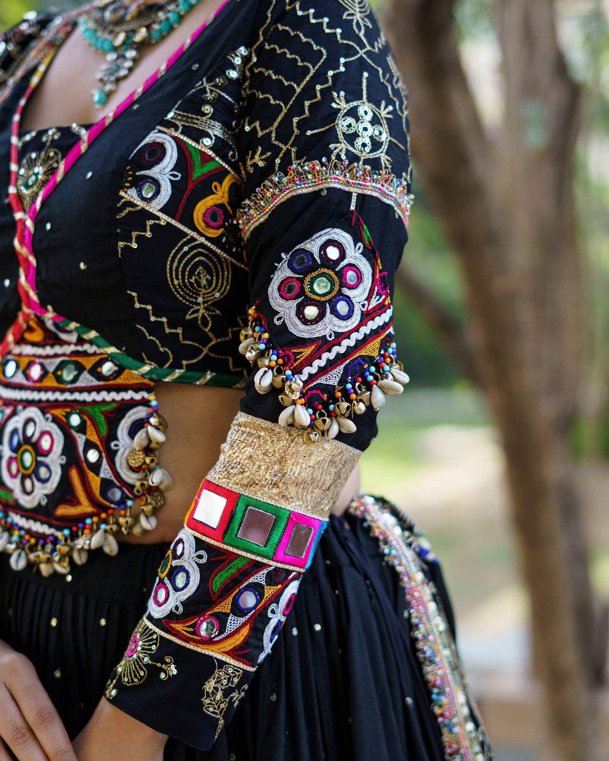 Detailed close-up of black blouse sleeve showcasing colorful mirror embroidery patches, gold cuff trim, and hanging cowrie shells