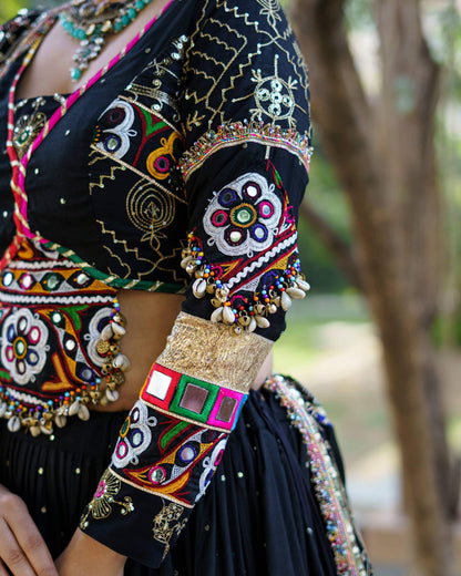 Detailed close-up of black blouse sleeve showcasing colorful mirror embroidery patches, gold cuff trim, and hanging cowrie shells