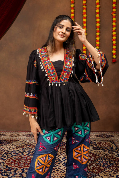Model wearing black V-neck peplum top with flared bell sleeves, shell fringe detailing, and embroidered denim pants
