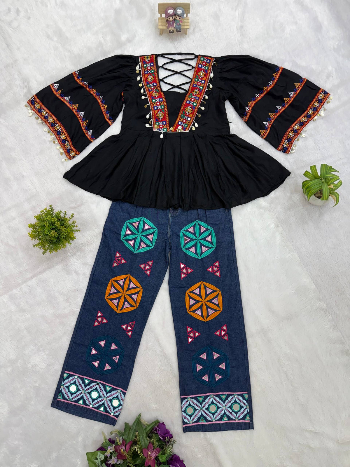 Flat lay view of black flare-sleeve peplum top with cowrie tassels and blue denim jeans featuring geometric mirror patches
