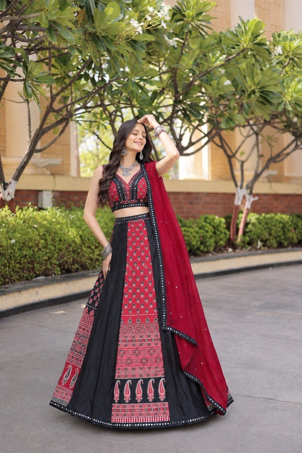 Model posing in an outdoor courtyard wearing a black and red flared lehenga skirt featuring traditional paisley motifs and silver oxidized jewelry.