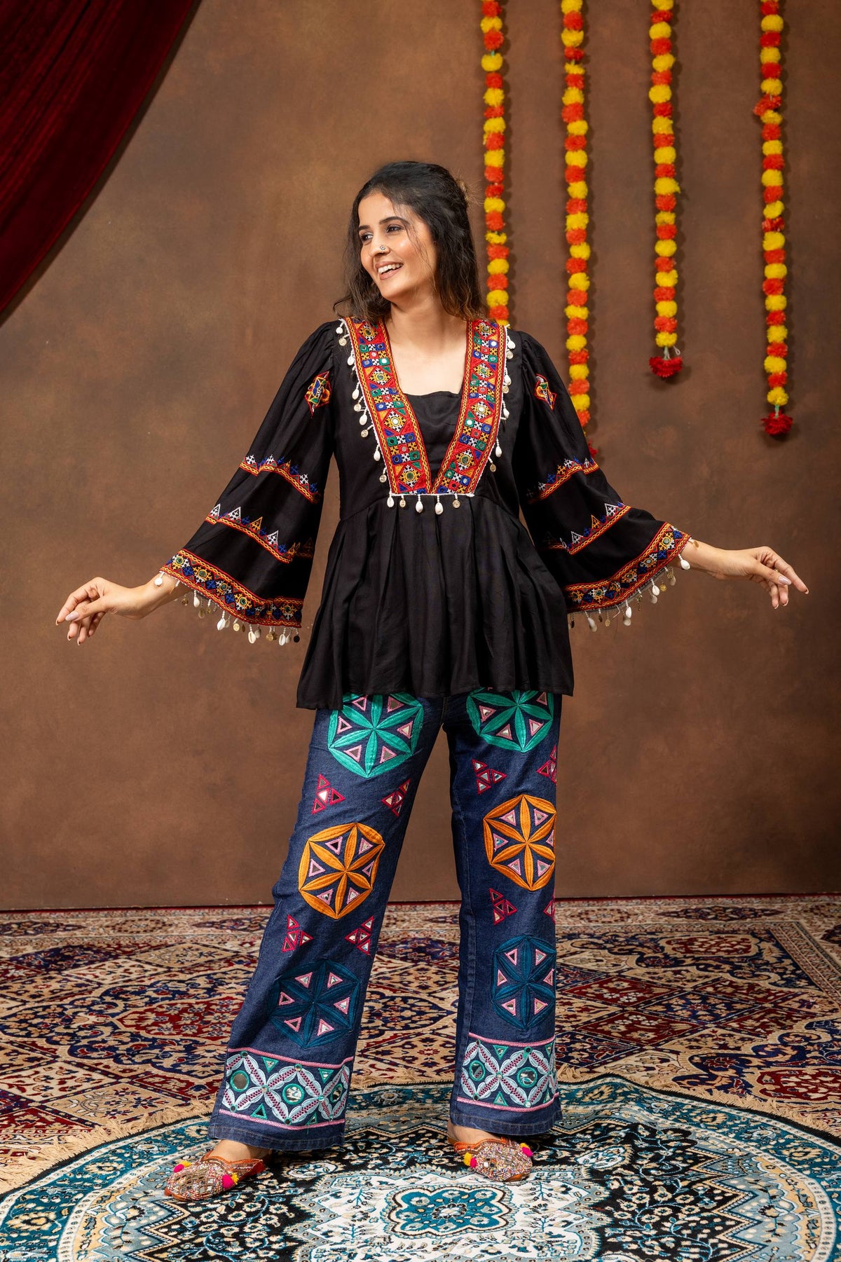 Full-length front pose of model wearing black embroidered tunic top with wide bell sleeves and wide-leg denim jeans