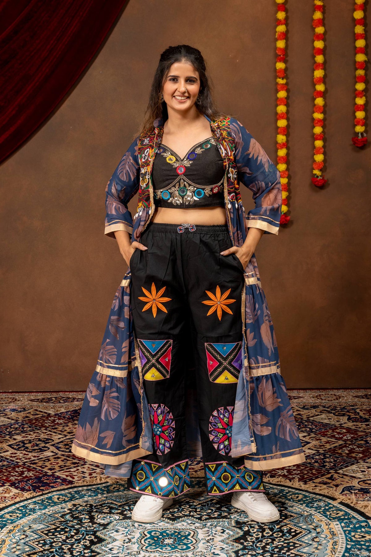 Model in a full-length pose wearing a blue floral print tiered long jacket, black embroidered crop top, and palazzo pants.