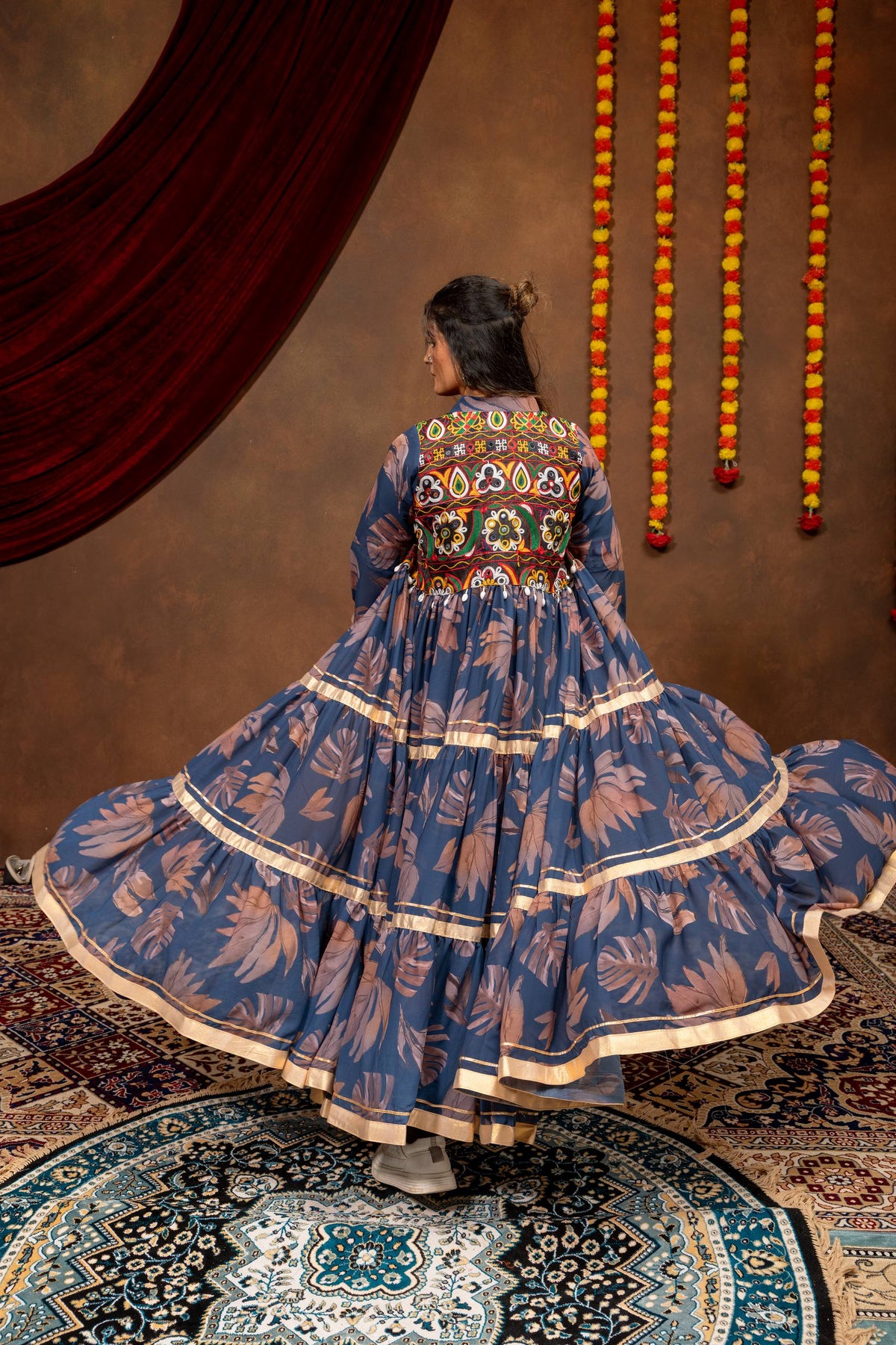 Rear view showing the circular tiered flare and colorful embroidered back yoke of a blue printed long jacket shrug.