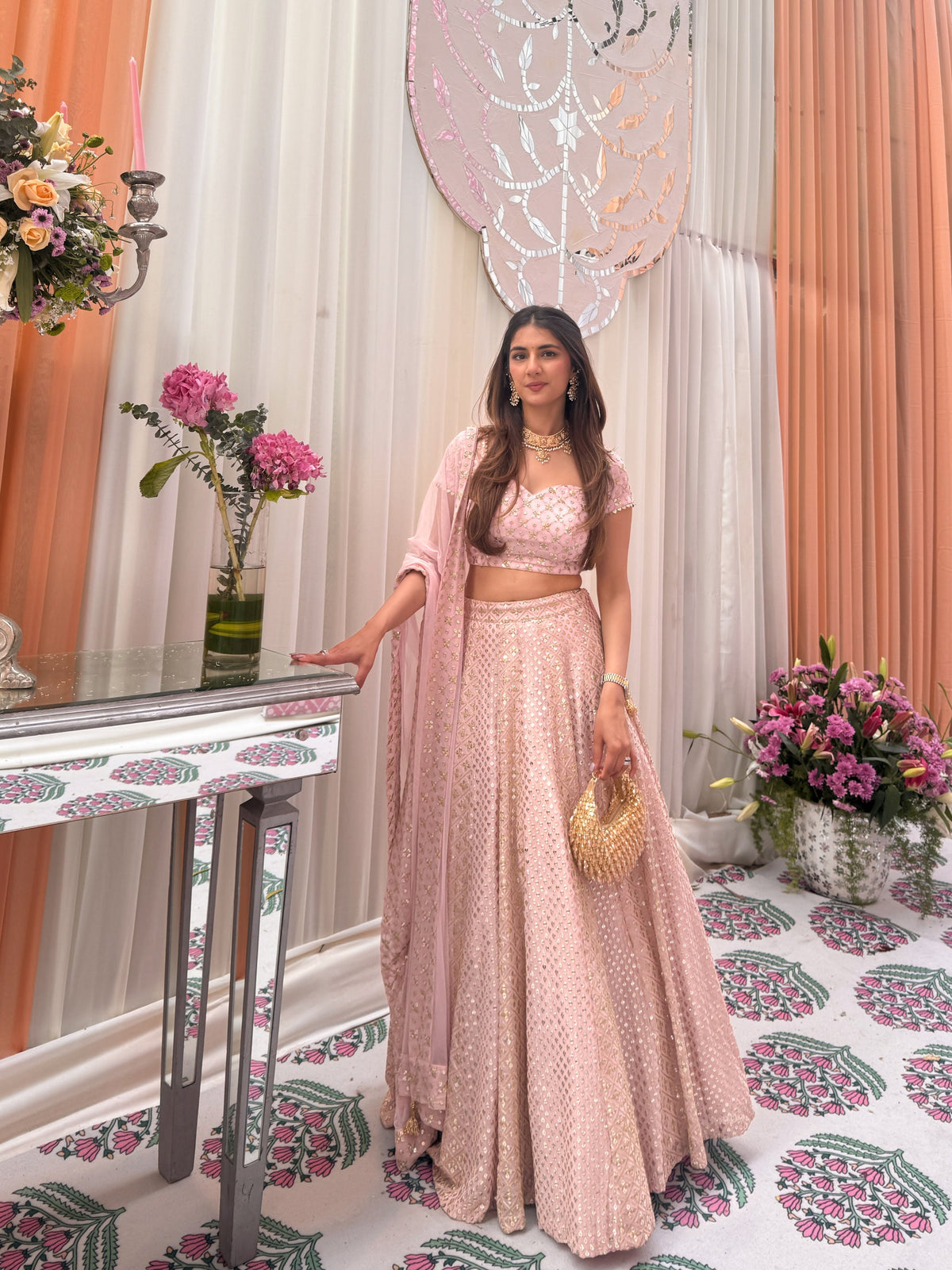 Standing full-length view of a model in a blush-pink flared lehenga, sweetheart-neck blouse, and sheer shoulder dupatta drape