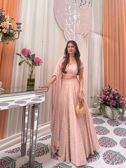Standing full-length view of a model in a blush-pink flared lehenga, sweetheart-neck blouse, and sheer shoulder dupatta drape