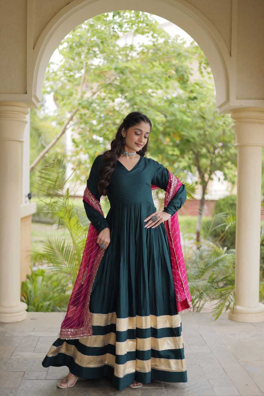 Full-length pose of a model wearing a bottle green V-neck anarkali gown with broad gold borders and a mirror-work pink dupatta.