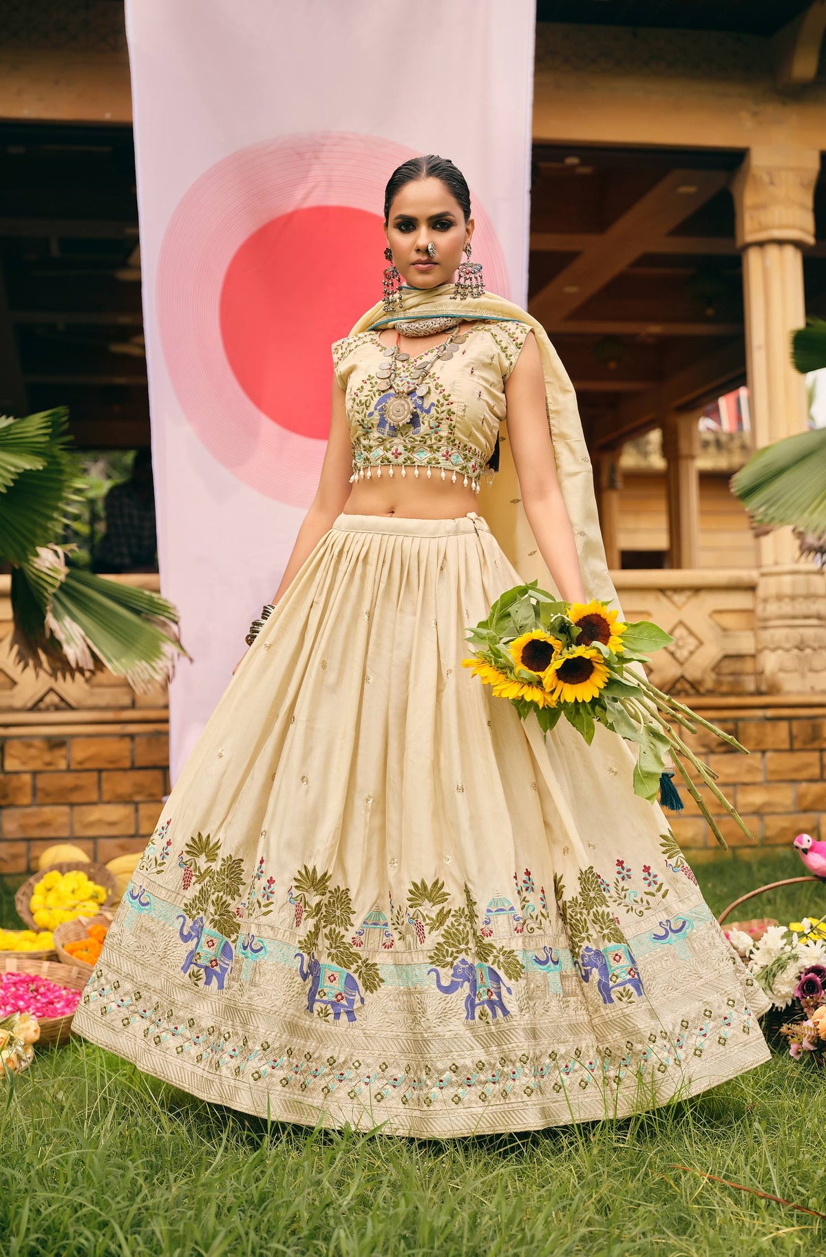 Full-length outdoor shot of model showcasing the wide embroidered hem flare of a cream lehenga choli holding yellow sunflowers