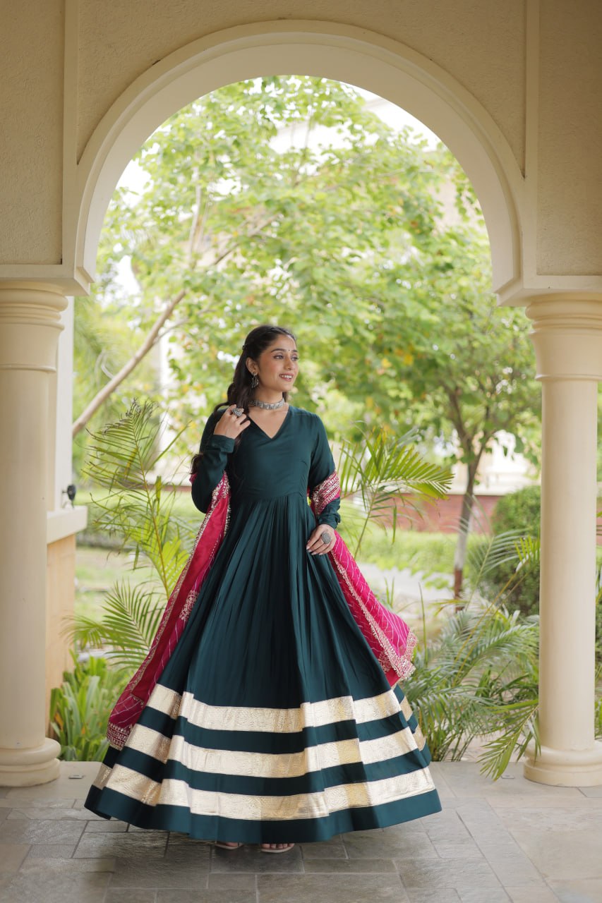 Outdoor full-length view of a model in a deep green flared anarkali dress highlighting dual wide gold zari hem stripes.