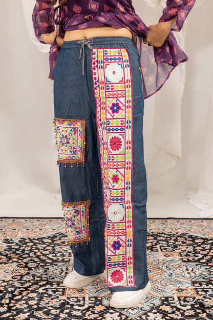 Side angle view of model wearing wide-leg denim trousers with colorful embroidered floral panels and mirror work patches