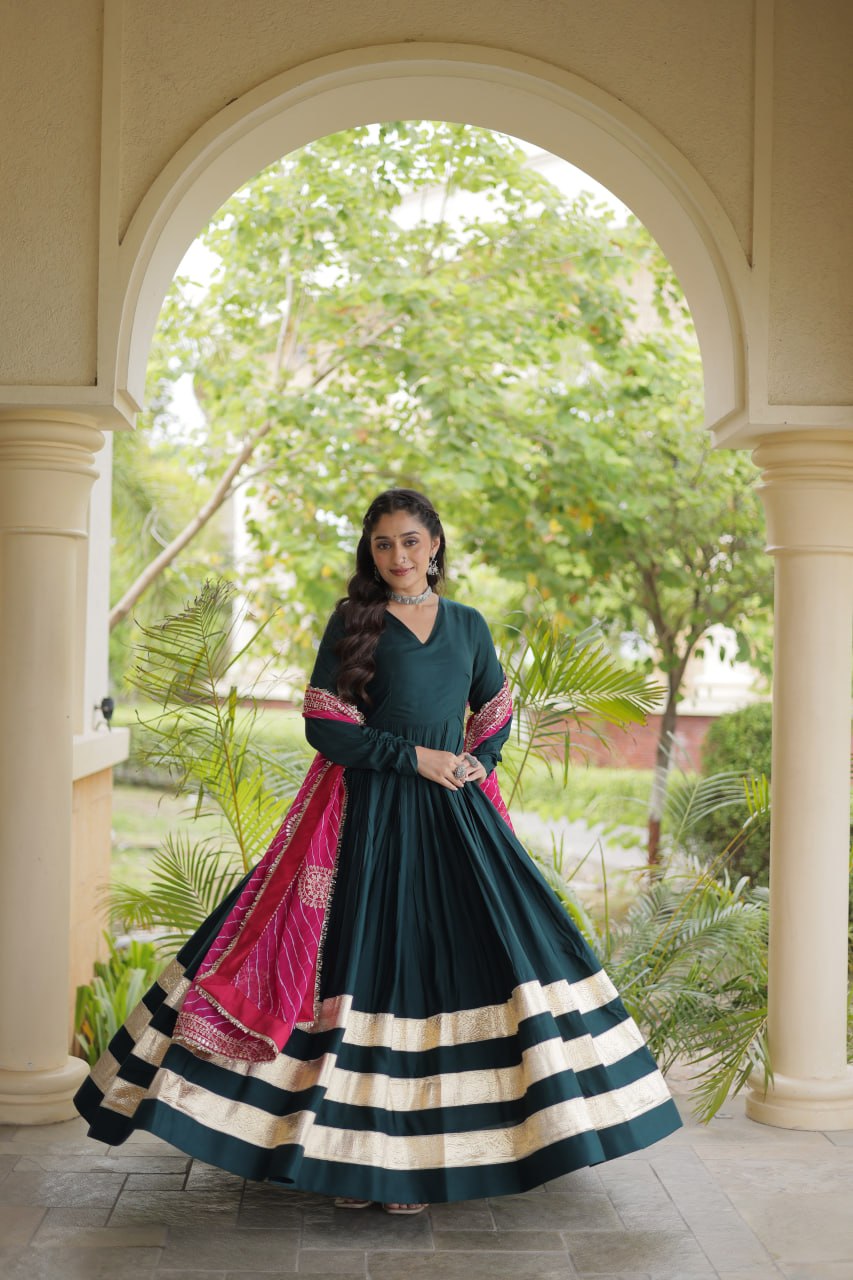Full-length shot capturing the wide flare of an emerald green tiered gown with bold gold border stripes and draped pink dupatta.