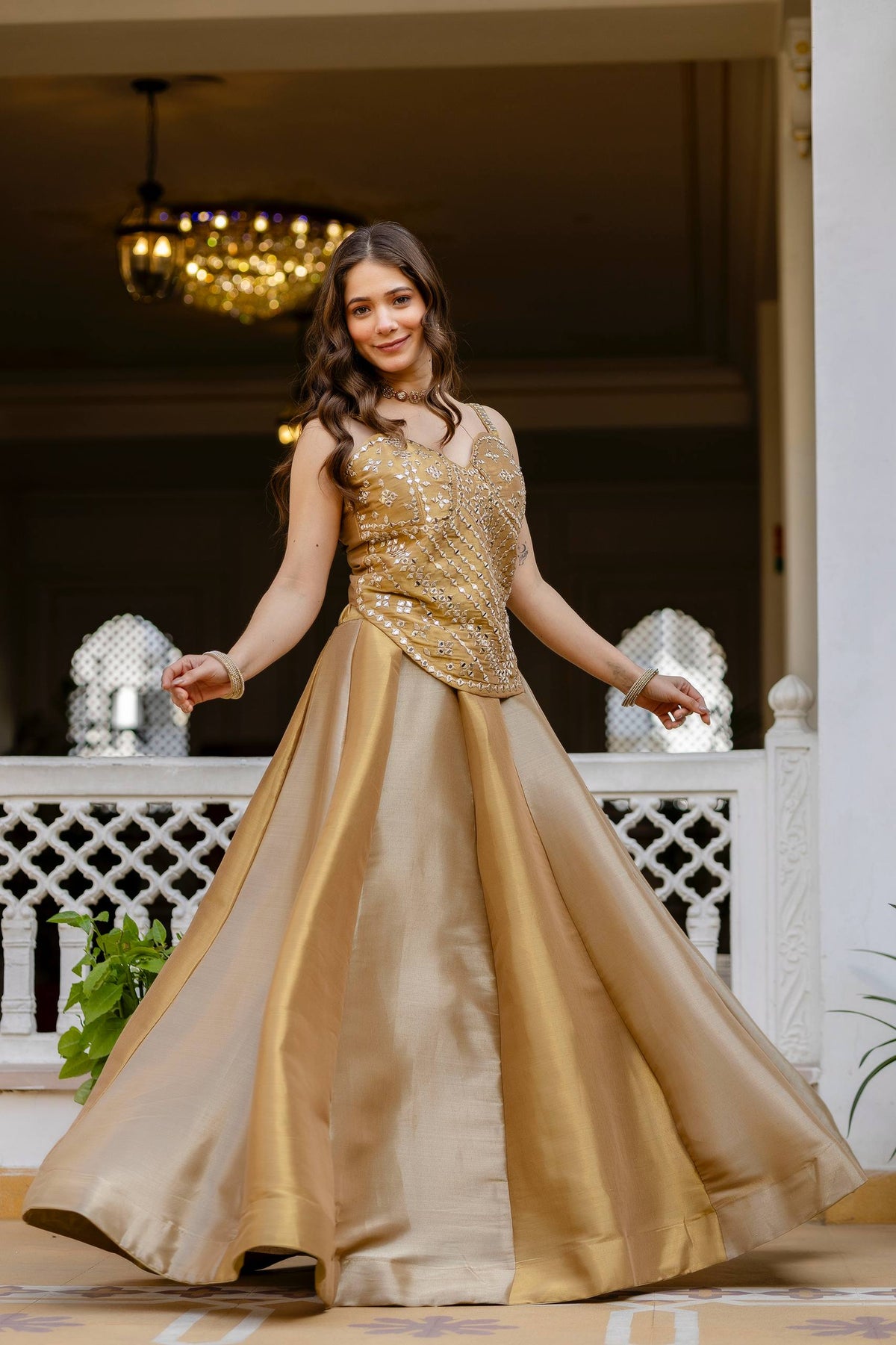 Full-length front standing pose of model in a wide flare gold panel lehenga and matching sleeveless mirror-work corset top
