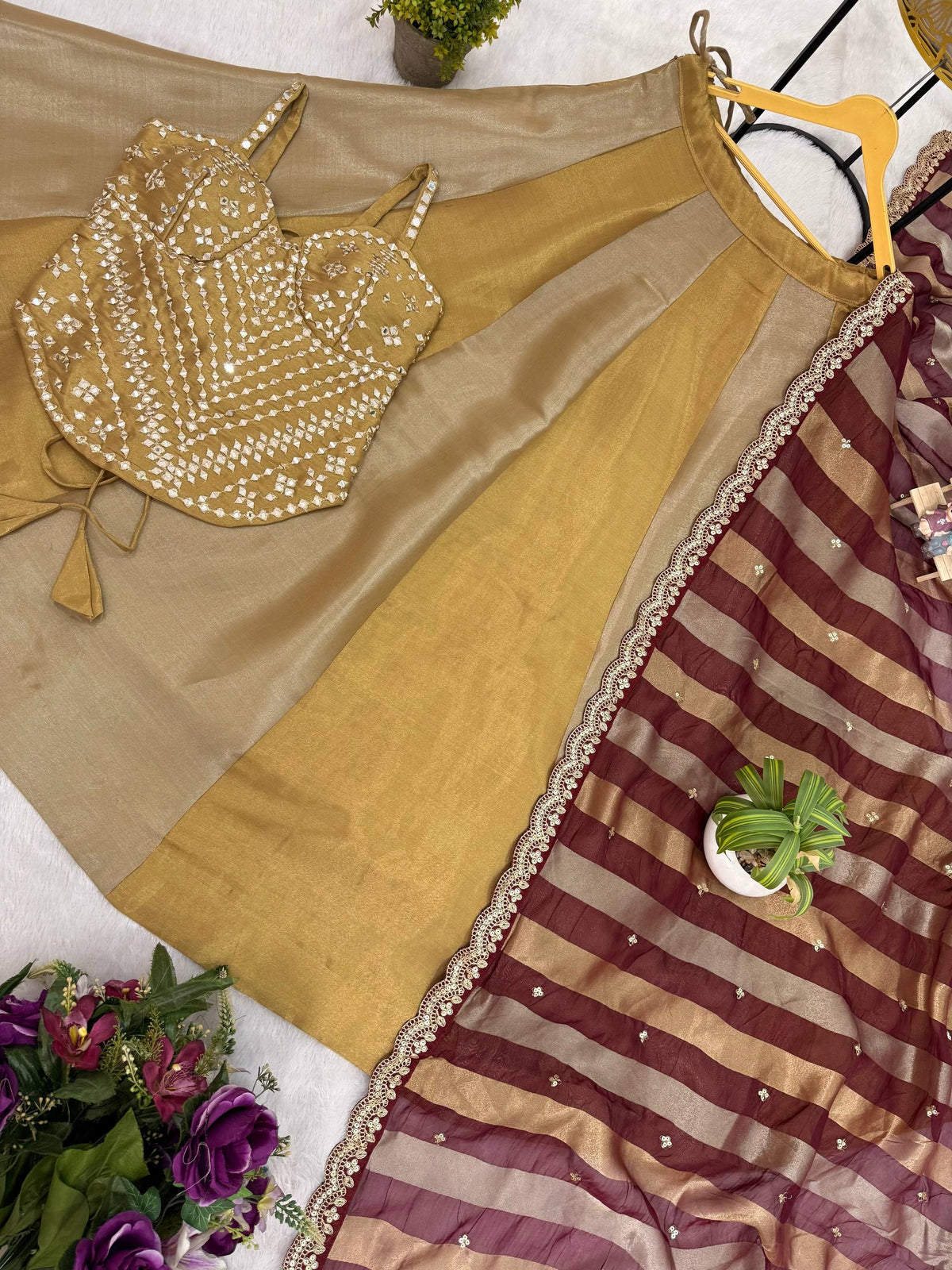 Flat lay of a dual-tone gold flared lehenga skirt, mirror embellished sleeveless corset blouse, and maroon-gold striped dupatta
