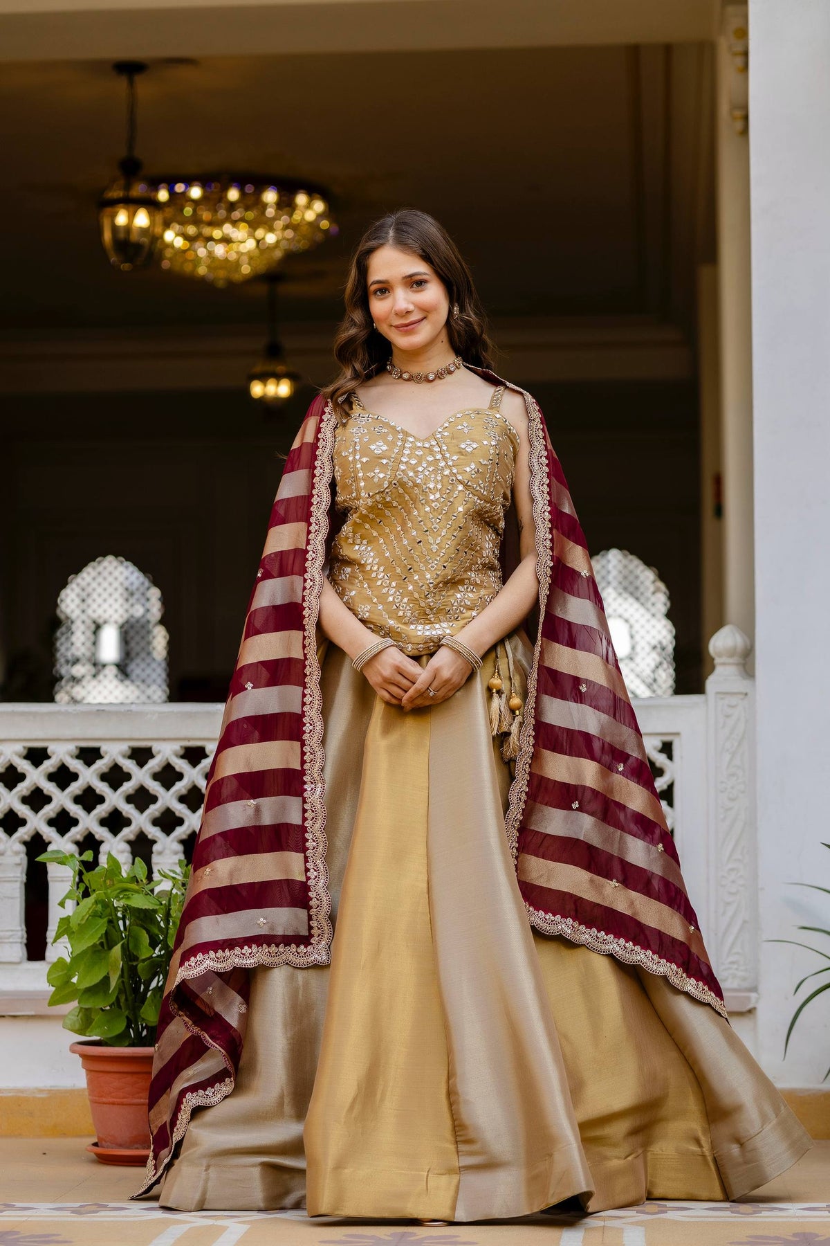 Full-length front view of model in a gold flared lehenga skirt and mirror-work corset blouse with a maroon striped dupatta draped over shoulders