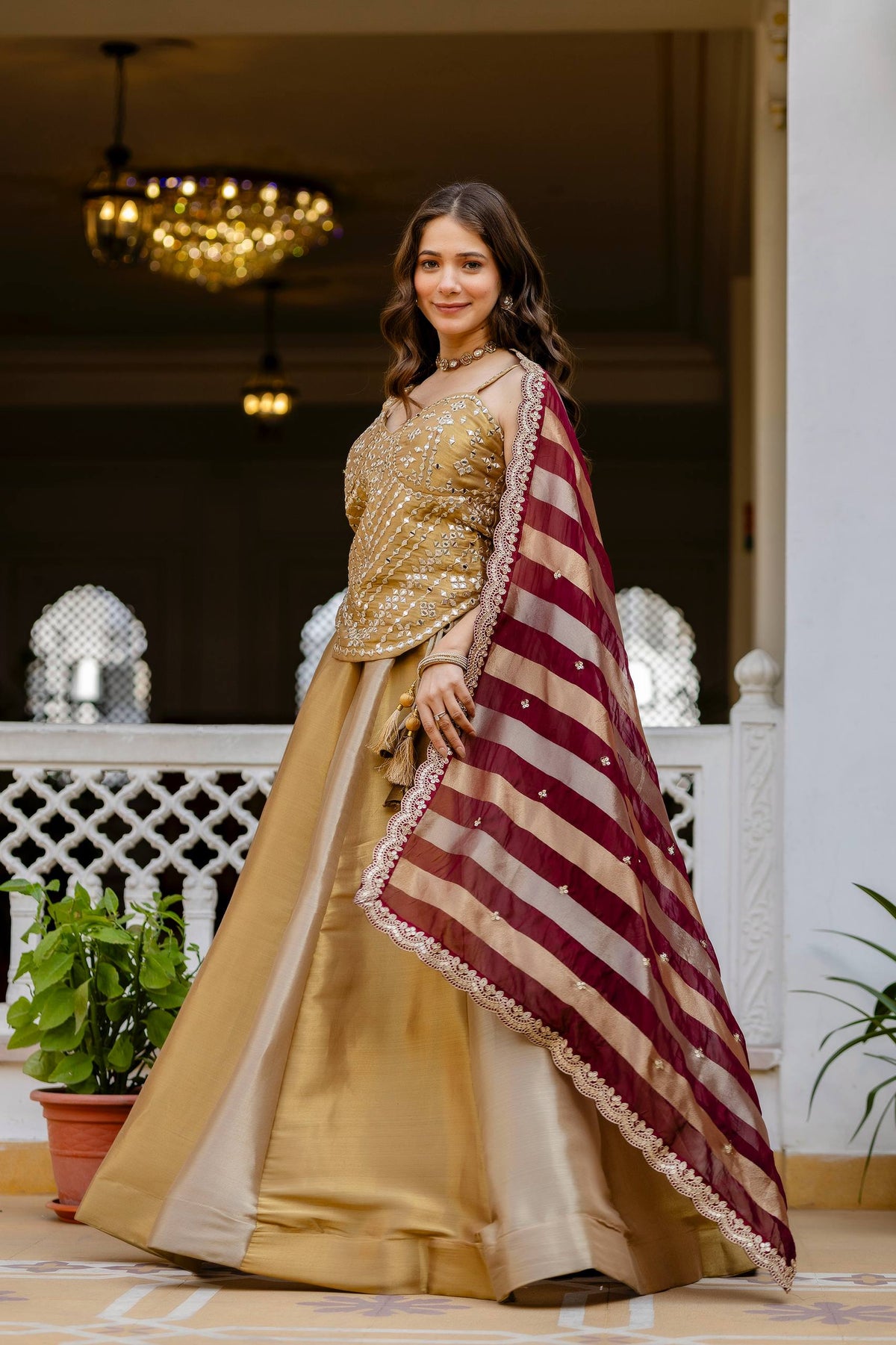 Side-angle full-length view of model in a gold panel flared lehenga styled with a maroon striped dupatta with scallop border