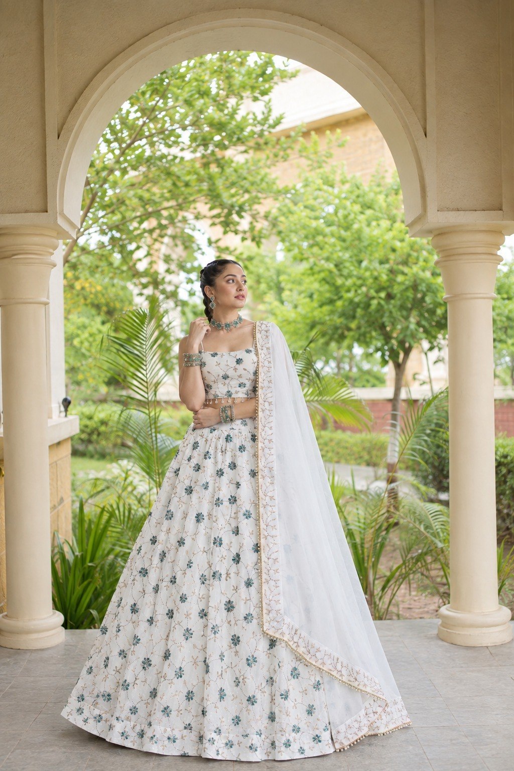 Full-length outdoor shot of a model wearing an ivory lehenga with allover green floral motifs, pearl waist drop accents, and sheer dupatta.