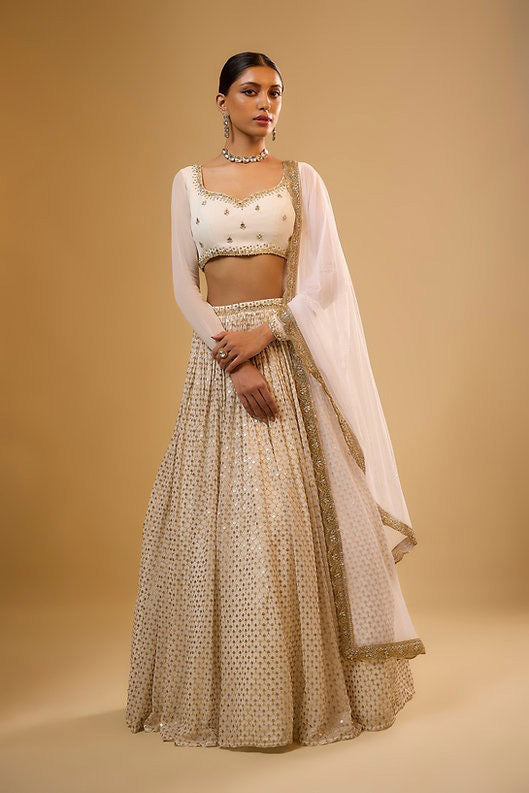 Full-length front view of a model wearing an ivory-white pleated lehenga with gold sequin embellishments, sweetheart-neck choli, and sheer dupatta.