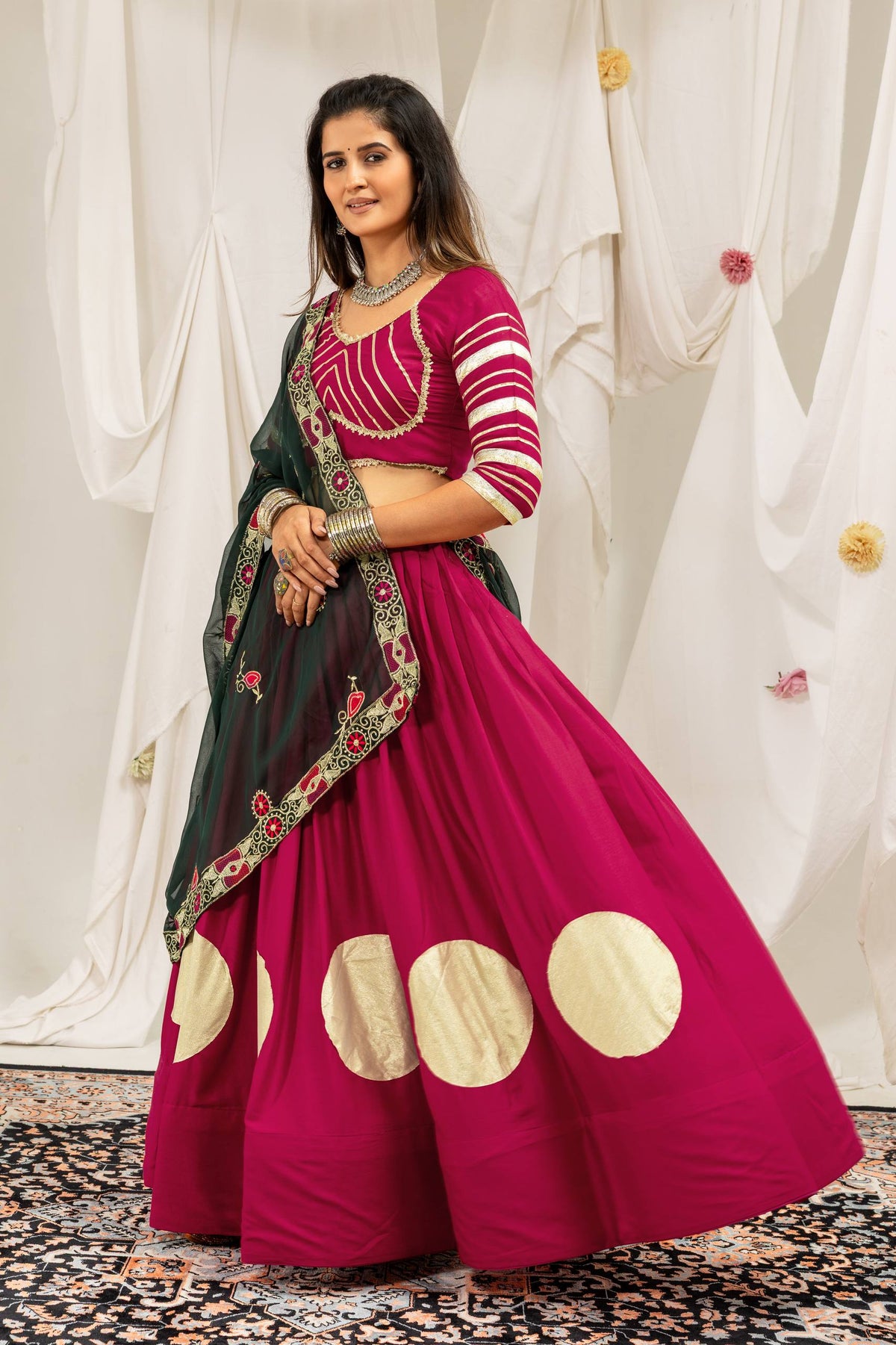 Model posing in a flared magenta lehenga with large gold foil circles and an elbow-sleeve choli with gold stripe detailing.