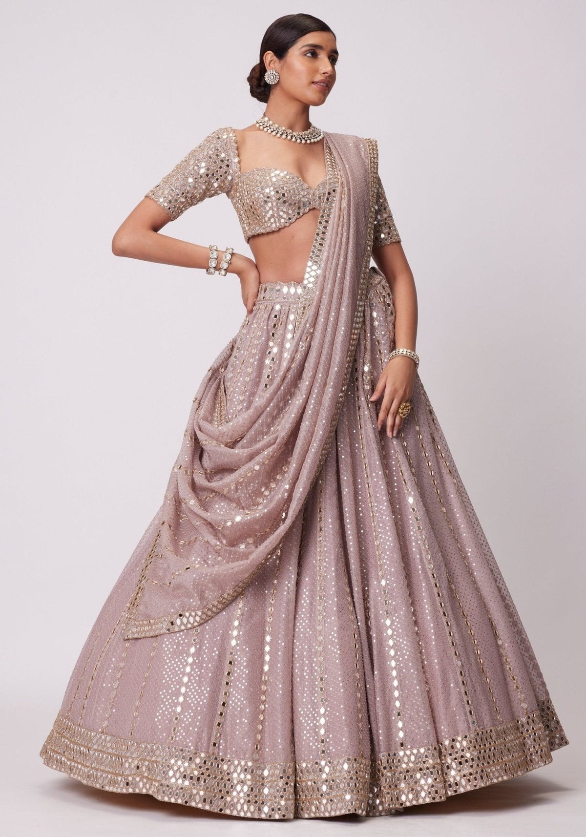 Side-angle full-length shot of a model wearing a mauve-pink mirror-embellished flared lehenga with cowled front dupatta drape