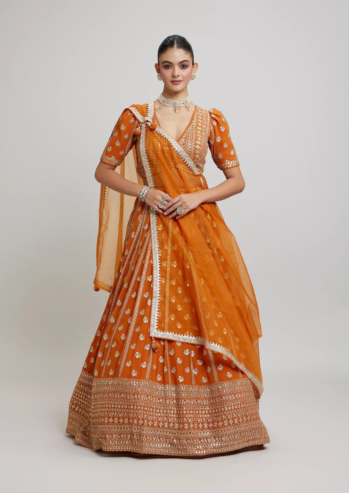 Full-length front view of a model wearing a mustard-orange lehenga choli with silver sequin embroidery and sheer organza dupatta.