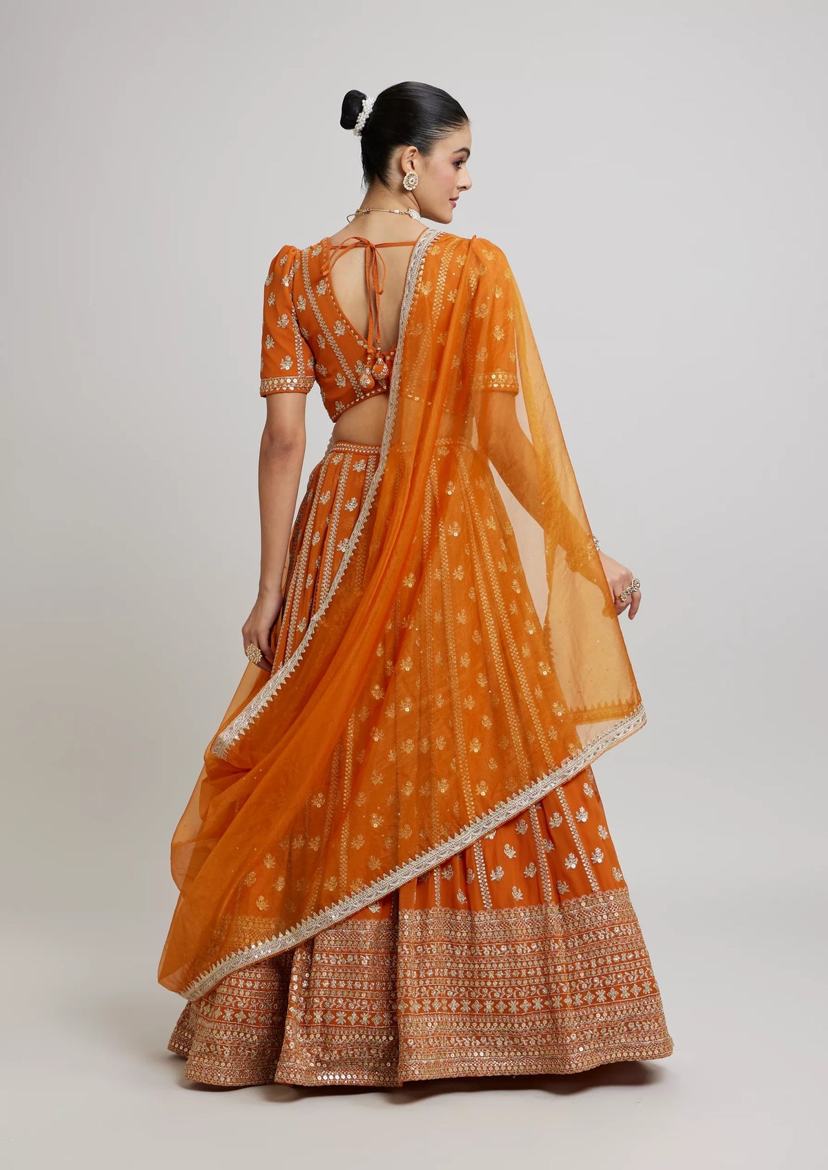 Full-length back view of a model in a mustard-orange flared lehenga and puff-sleeve blouse with tie-up back dori.
