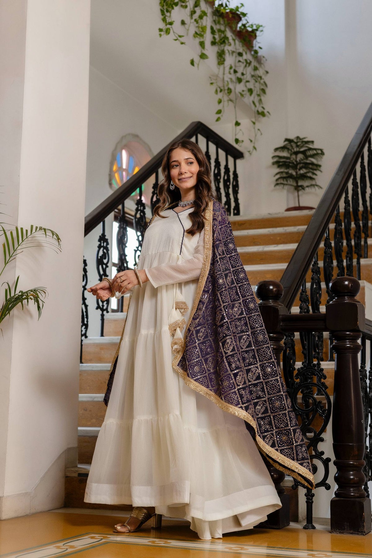 Standing side-angle view of model wearing an off-white tiered anarkali suit with a draped purple and gold printed dupatta