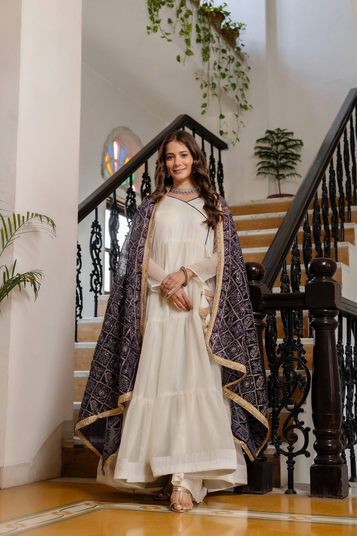 Full-length front view of model in an off-white tiered anarkali dress with deep purple gold-bordered dupatta draped across shoulders