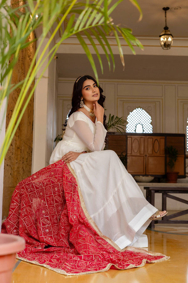 Model seated wearing an off-white tiered anarkali dress with a red decorative dupatta spread on the floor