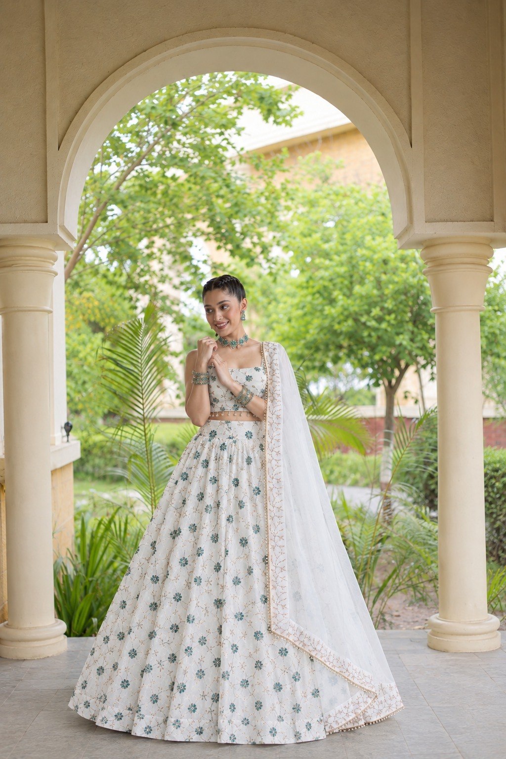 Standing full-length front view of a model under an archway wearing an off-white flared lehenga adorned with green floral thread embroidery.