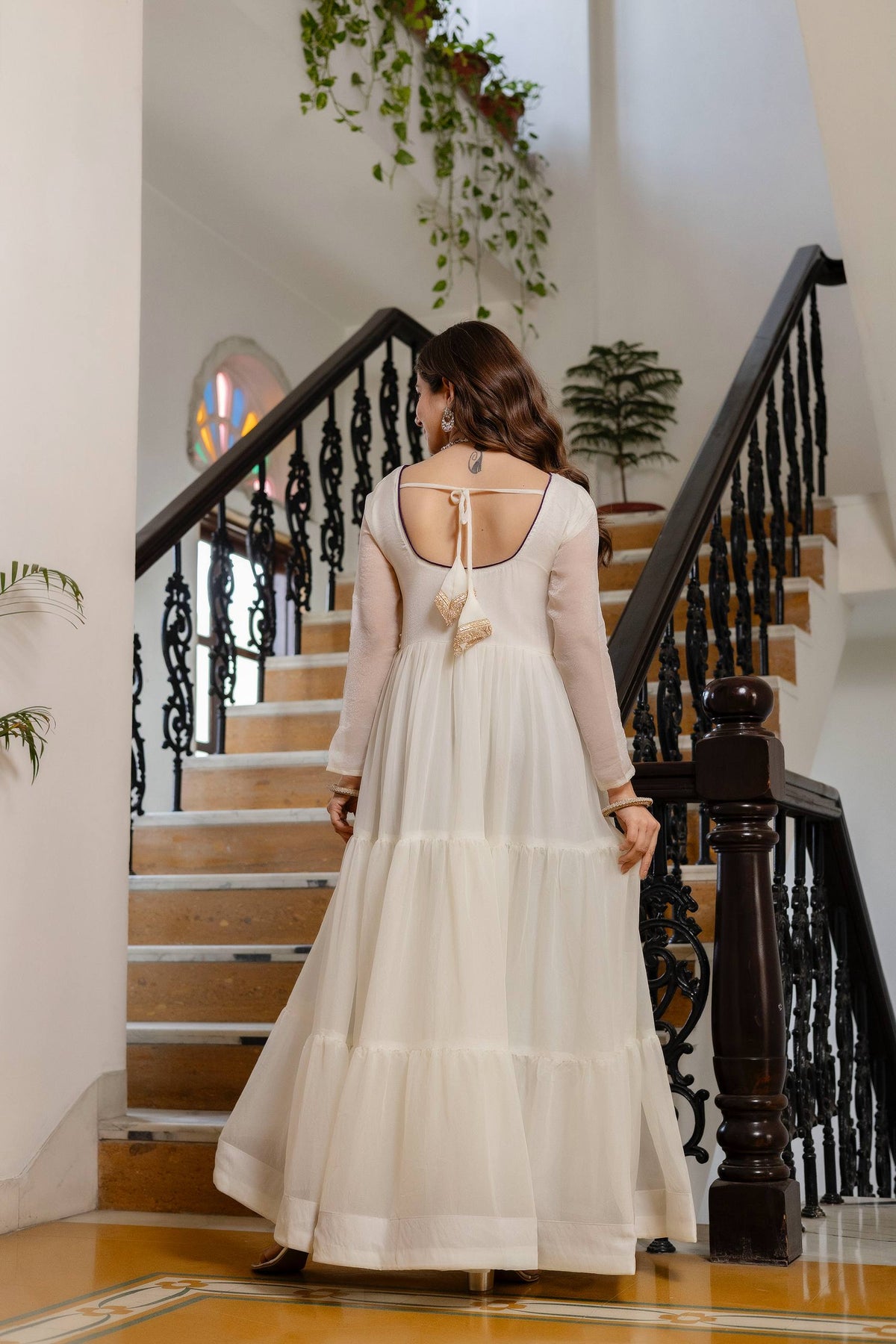Full-length back view of an off-white tiered anarkali suit showing deep round back neck with tie-up latkan tassels