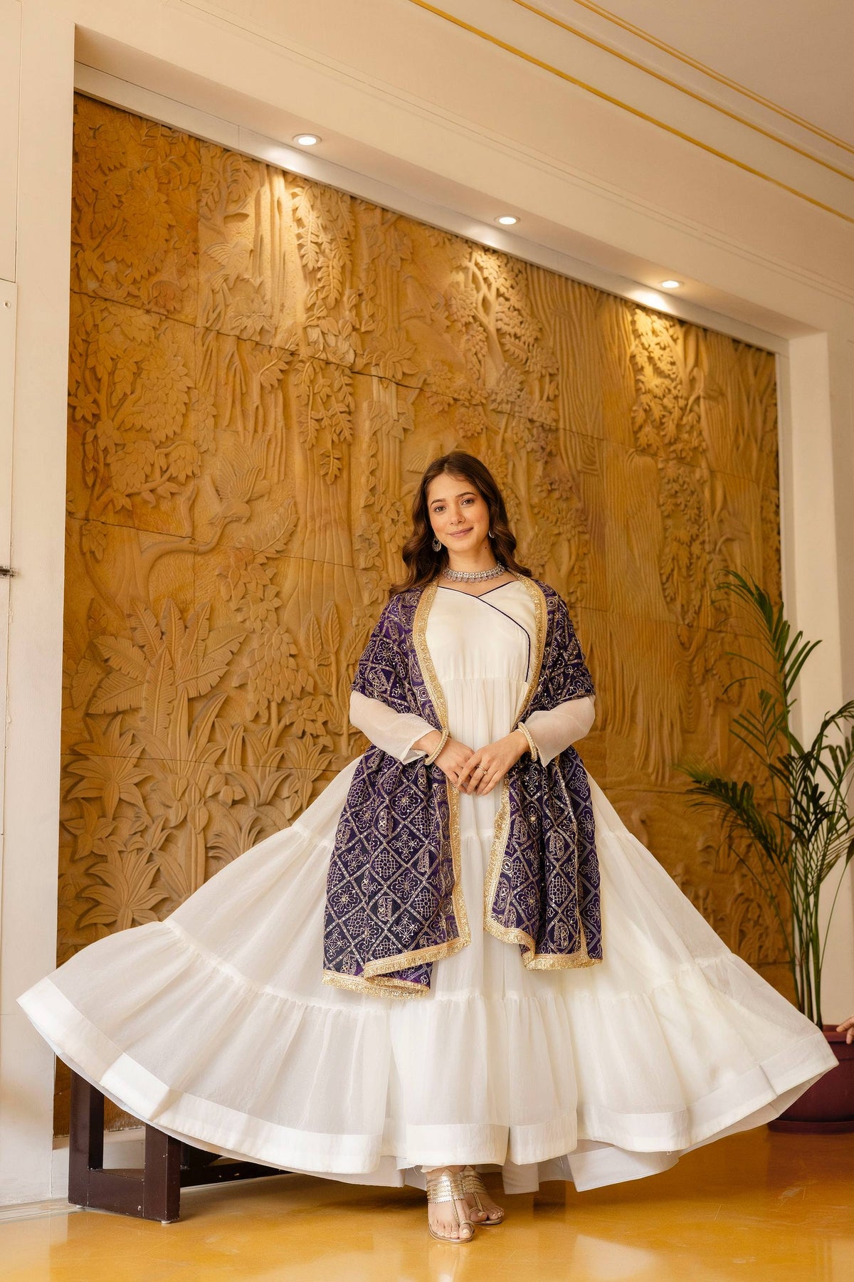 Full-length shot of model showcasing the wide tiered flare of an off-white anarkali suit with draped deep purple dupatta