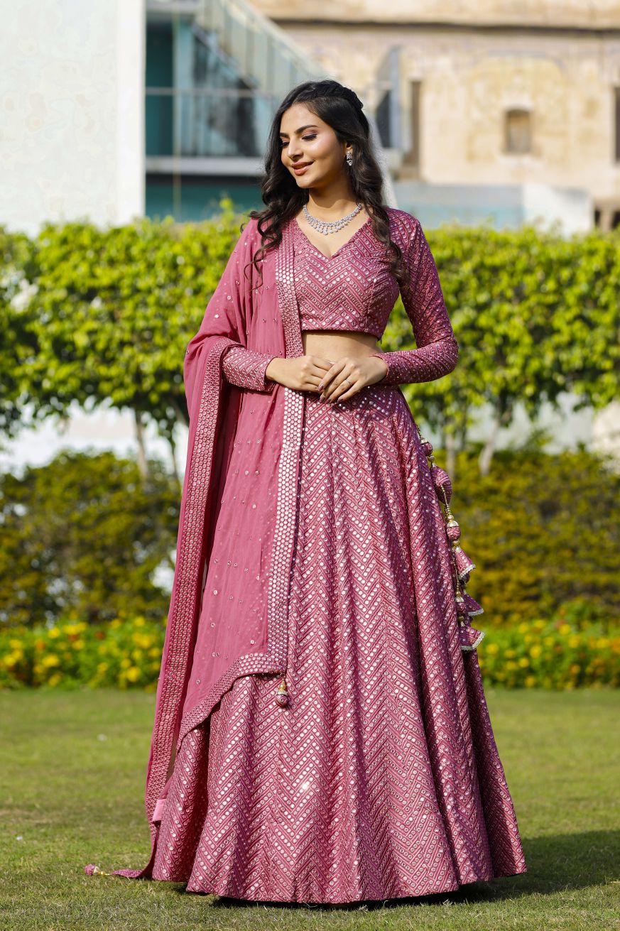 Model posing in a sunny garden setting wearing an onion-pink mirror-work lehenga choli set with side-draped dupatta.