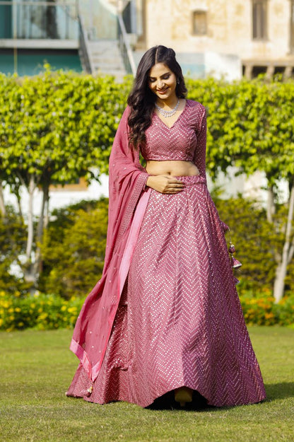 Full-length front view of a model in a lawn setting wearing an onion-pink lehenga with chevron mirror embroidery and a full-sleeve blouse.
