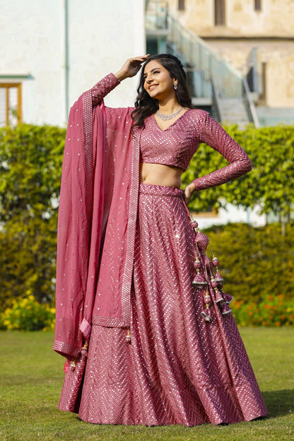 Standing full-length front view of a model wearing an onion-pink lehenga with decorative latkan tassels and sheer matching dupatta.