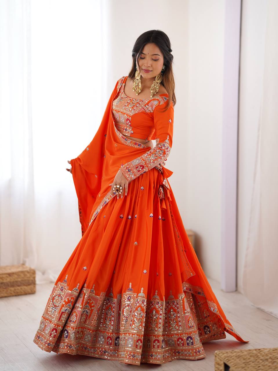 Model posing in an orange flared lehenga choli with full-sleeve blouse and wide embroidered skirt border