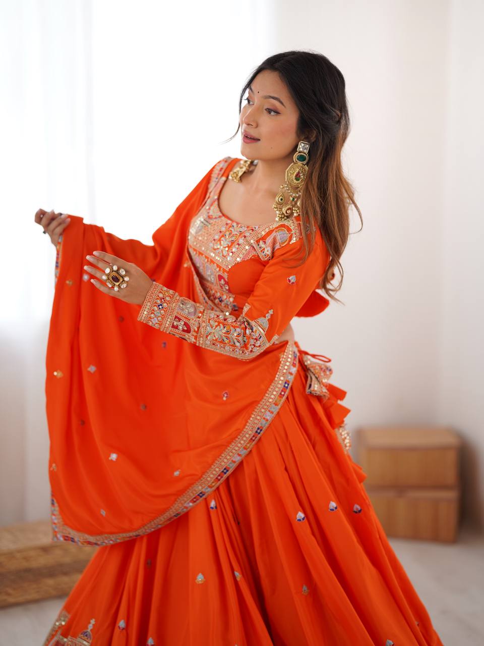 Model wearing an orange full-sleeve embroidered blouse and draped dupatta with colorful mirror-work border