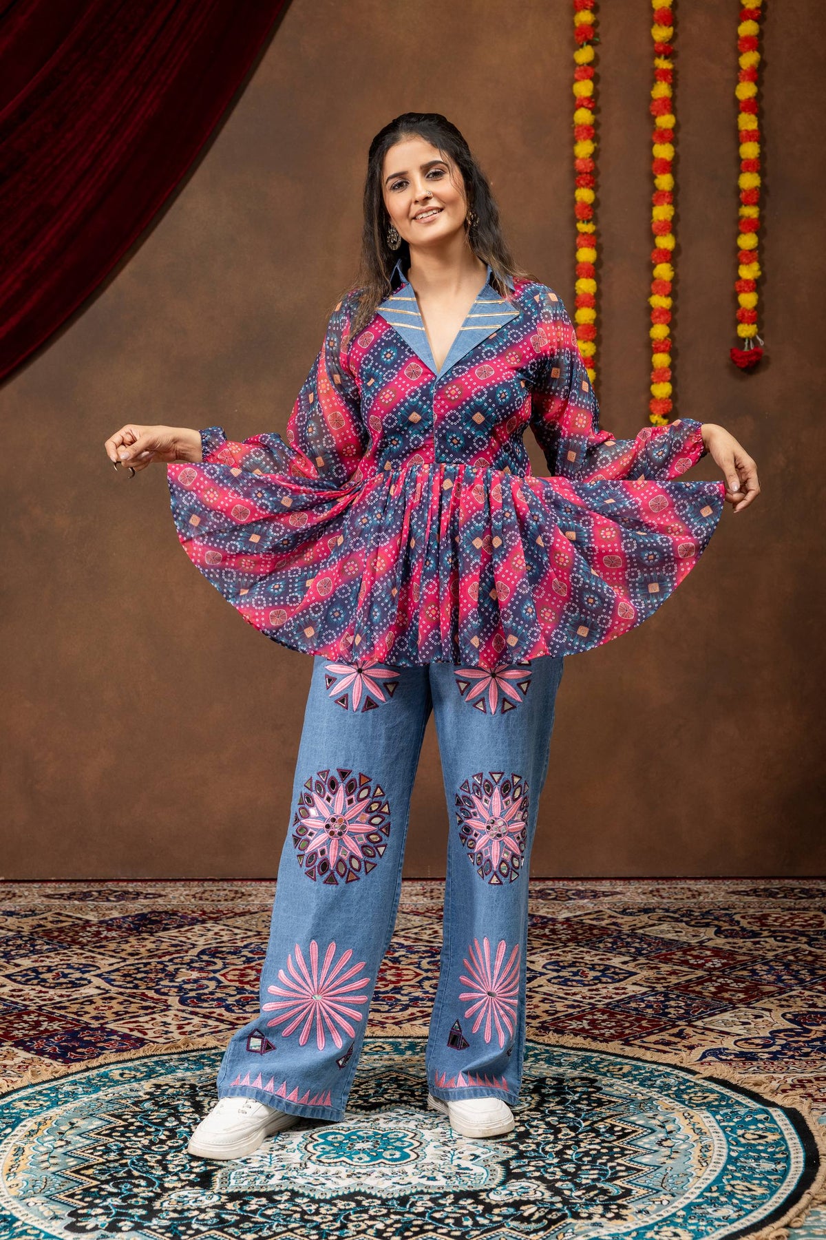 Model spreading hem of pink and blue bandhani printed flared peplum top paired with wide-leg mirror work denim jeans