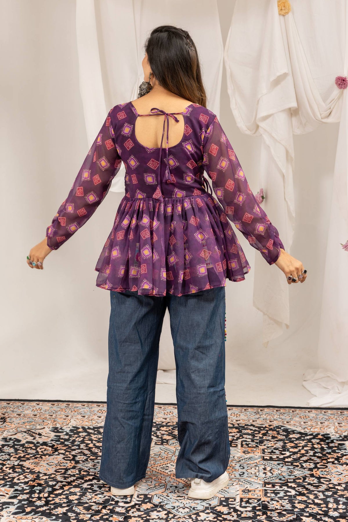 Back view of model wearing purple printed peplum top with tie-up back neckline and wide-leg denim jeans