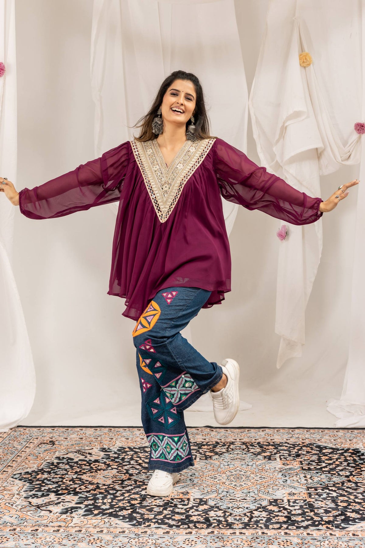 Model in purple pleated v-neck tunic with embroidered neckline and geometric embroidered blue denim jeans