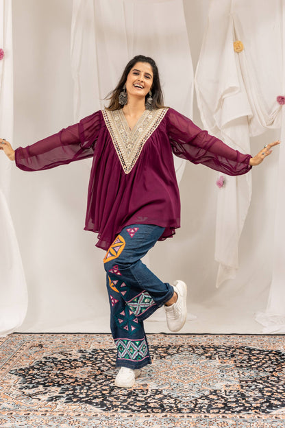 Model in purple pleated v-neck tunic with embroidered neckline and geometric embroidered blue denim jeans