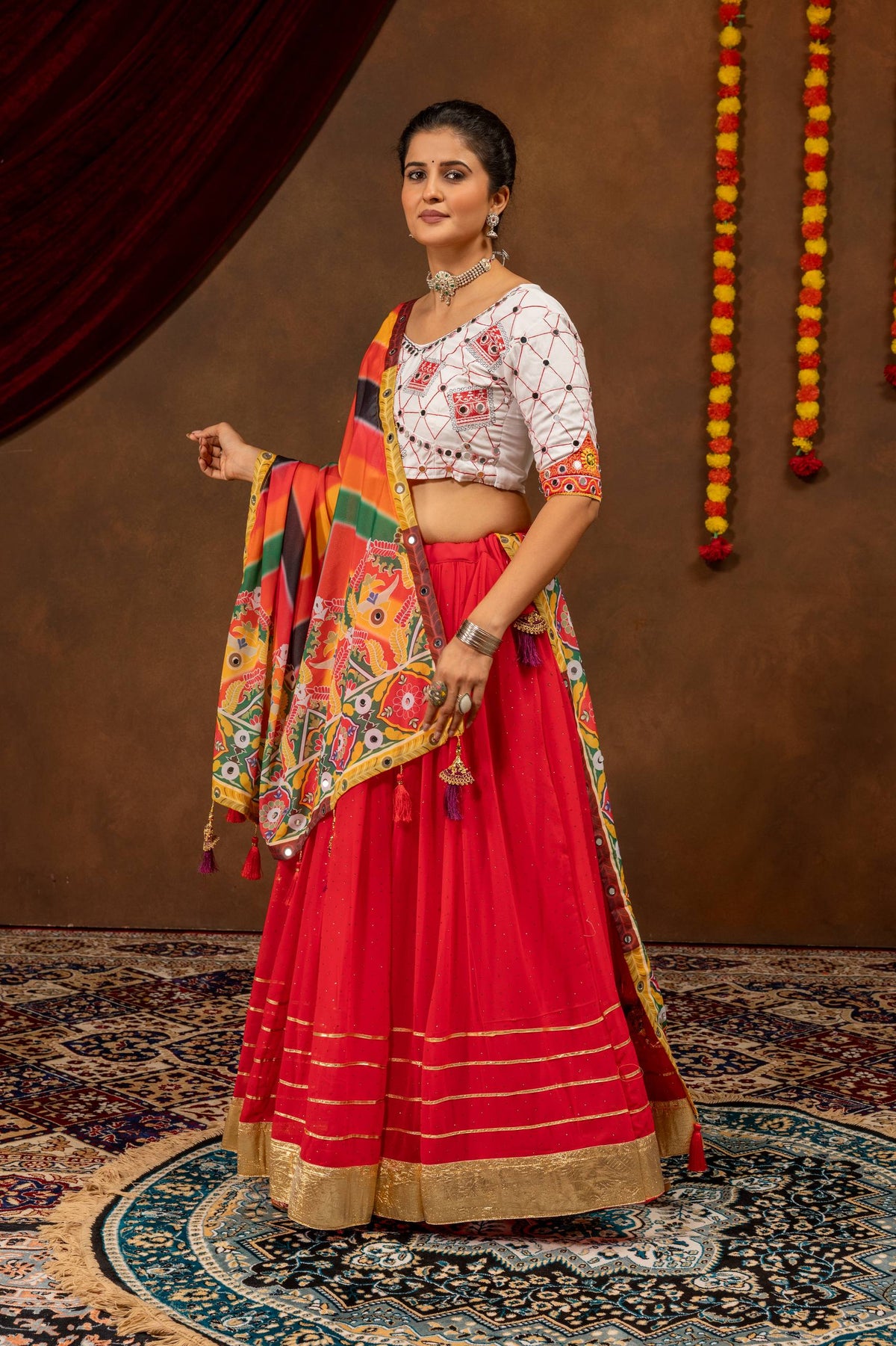 Three-quarter standing pose of a model dressed in a red lehenga with broad gold hemline borders and a white embroidered choli.