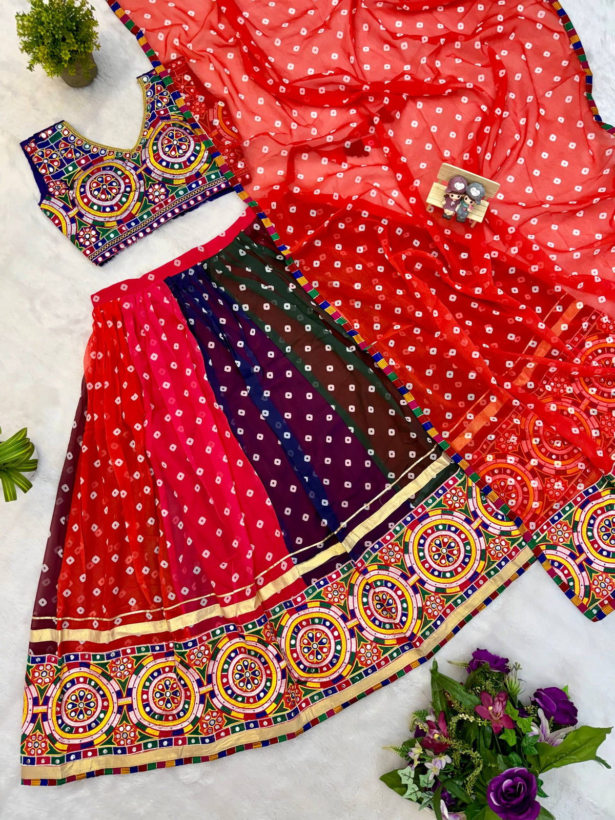 Flat lay view of red and multicolor panelled bandhani lehenga, Kutchi mirror work blouse, and red bandhani dupatta