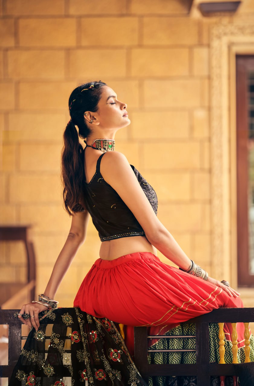 Side-profile pose of model seated on a wooden bench in a pleated red lehenga skirt with gold gota lines and a black sleeveless blouse